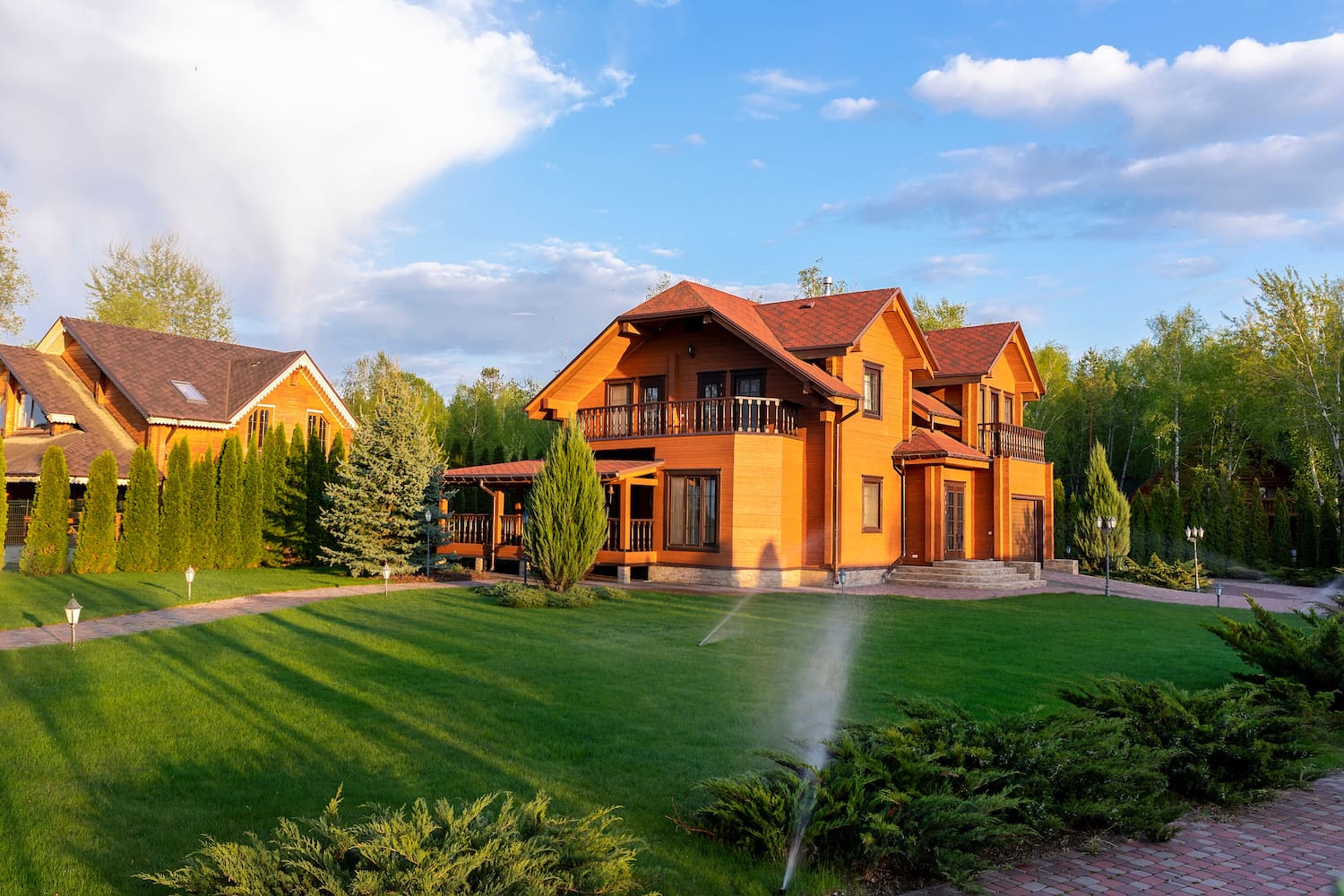 Modern wooden eco house villa facade luxury big house. Timber cottage with with green lawn water sprinkler, paved footpath and blue sky background. Landscaping design, garden watering and maintenance